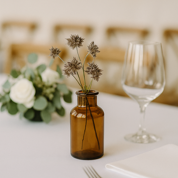 Table setting with a brown bottle, flowers, and a glass on a white tablecloth.